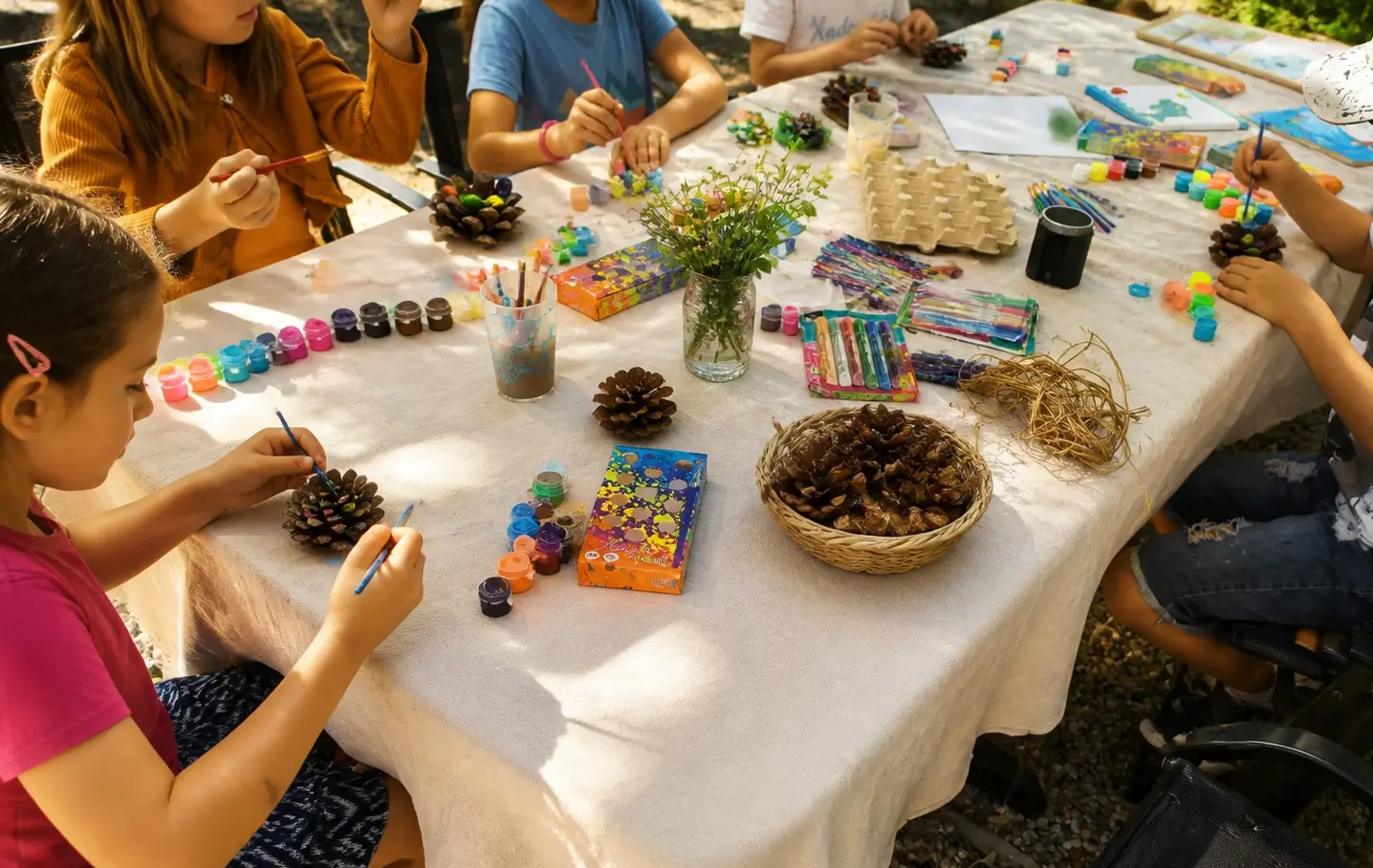 Children painting pine cones around a cloth-covered table outdoors in dappled light